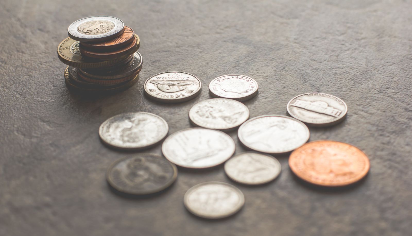 A stack of coins beside assorted loose coins on a grey stone surface.